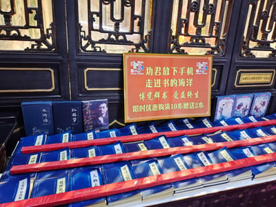 图片展示了一个传统风格的书籍促销场景。背景是一扇具有中国古典特色的黑色木门，门上有精美的雕花装饰。门前摆放着一个书摊，整齐地陈列着蓝色封面的书籍，包括《水浒传》《红楼梦》等经典著作。书摊上方有一个醒目的红色标语牌，上面写着“劝君放下手机 走进书的海洋 博览群书 受益终生 限时优惠购满10本赠送2本”，倡导人们减少手机使用，多读书。整个场景融合了传统文化元素与现代阅读推广理念，营造出一种鼓励阅读的氛围。