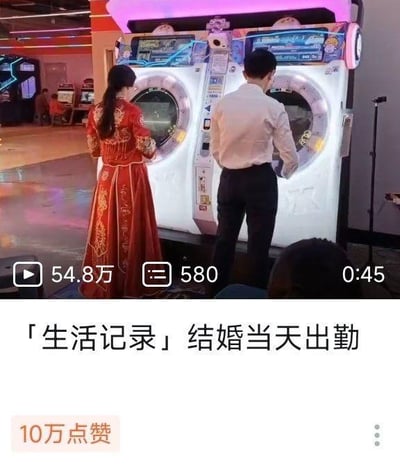 这是一段短视频的截图，画面中一位身着中式红色秀禾服的新娘与一位穿着衬衫西裤的新郎，正站在游戏厅的双人游戏机前专注地操作机器，看起来正在游玩游戏。视频下方标注的标题为「生活记录」结婚当天出勤，这里的“出勤”是双关用法，原本指按时到工作岗位上班，此处调侃新人在结婚当天没有按常规待在婚礼相关流程里，反而像“上班出勤”一样来到游戏厅打游戏，充满了反差感，十分有趣。视频的播放数据显示有54.8万次播放、580条评论、时长45秒，还收获了10万点赞。