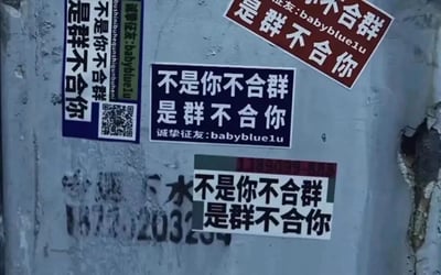 这张图片展示了张贴在粗糙且带有磨损痕迹墙面上的四张标语贴纸，四张贴纸设计风格各异，但核心标语完全一致——「不是你不合群，是群不合你」。其中三张贴纸附带「诚挚征友:babyblue1u」的信息，还有一张仅保留核心标语：左上角是蓝底白字的贴纸，附带二维码与征友信息；右上角是红底白字的贴纸，同样标注征友信息；中间是黑底白字的贴纸，清晰印有征友内容；下方是灰黑配色的贴纸，仅显示核心标语。墙面下方还残留着模糊的「下水2032」字样的标记，整体场景呈现出街头随意张贴的物料特征，标语以反向共情的角度，慰藉在社交中感到格格不入的人群，同时借此征友寻找同频伙伴。
