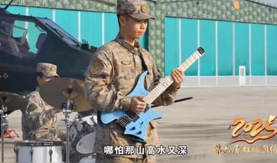 画面中呈现了两名身着荒漠迷彩军装的军人组成的小型乐队，前景的军人手持一把蓝色无头电吉他，神情专注地进行演奏；背景的军人则在架子鼓前投入地敲击鼓面。他们身处一处开阔的室外场地，背景中可以看到一架军用直升机和带有绿色挡板的建筑，场地看起来像是一处军用机场坪。画面右下角带有2025蛇年的艺术字图案以及“第六届雄师网络”的字样，底部配有白色字幕“哪怕那山高水又深”，整体场景展现了军营文化生活中充满活力的一面，打破了人们对军人的刻板印象，体现了军人多才多艺的特质。