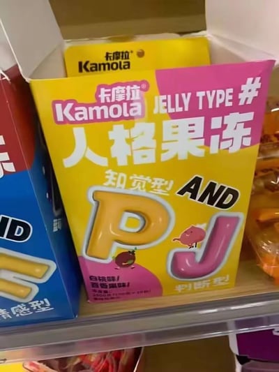 超市货架上的卡摩拉（Kamola）人格果冻包装盒，主体为黄色和粉色配色。包装正面醒目印有“人格果冻”大字，副标题为“知觉型 AND 判断型”，并以大号字母“P”和“J”突出展示，对应MBTI人格类型中的知觉（Perceiving）与判断（Judging）维度。下方标注口味为“白桃味/百香果味”，整体设计将心理学概念与零食结合，充满创意和趣味性。