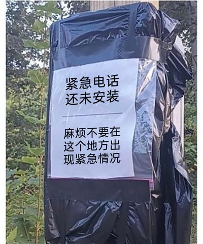 这是一张户外实拍的静态照片，画面主体是一个被黑色塑料布包裹的待安装紧急电话设施，设施表面贴着一张白色告示。告示分为上下两部分，上方文字为“紧急电话还未安装”，下方文字为“麻烦不要在这个地方出现紧急情况”，以一种诙谐自嘲的语气告知路人此处暂时无法提供紧急电话服务，用轻松的方式化解了设施未安装完成的尴尬，背景是户外的绿植环境，整体氛围搞笑有趣。