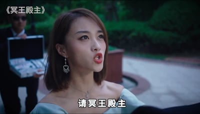 图片为网络小说《冥王殿主》的推广广告截图。画面中一位身着浅蓝色露肩连衣裙的女性，佩戴心形耳环和项链，短发，表情带着期待望向斜上方。背景中一名黑衣男子手持打开的公文包，站在绿植环绕的户外石板路上。左上角有标题《冥王殿主》，底部字幕显示“请冥王殿主”。该广告延续了“龙王赘婿”系列的“卑微请求-身份揭露”叙事模式，是2020年后流行的赘婿逆袭类网文广告典型风格。