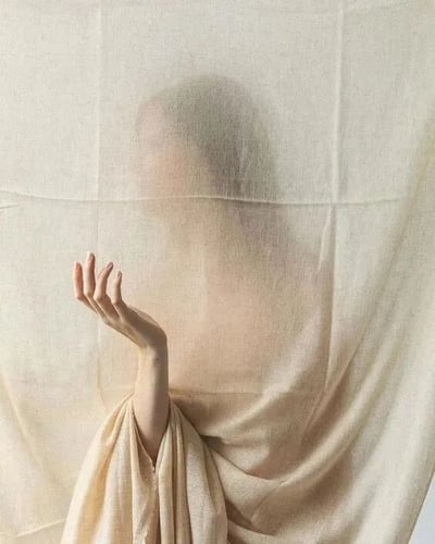 一张艺术摄影作品，画面中一位人物站在半透明的米色纱帘后，面部被布料朦胧遮盖，无法清晰辨认五官。人物伸出一只手，手指微微弯曲，轻柔地触碰着纱帘表面。整体色调为温暖的米白色，光线柔和，营造出一种神秘、宁静而富有诗意的氛围。布料的纹理和褶皱清晰可见，与人物模糊的轮廓形成鲜明对比，突出了触觉与视觉的微妙关系。