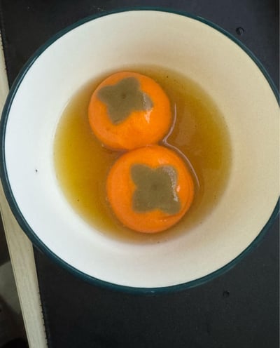 一张俯拍的食物照片，展示了一个白色内底、蓝色边缘的碗，碗中装有淡黄色液体，液体中浸泡着两个完整的橘子。每个橘子表面都有一个明显的深褐色星形印记，印记形状规则，位于橘子顶部。碗放置在深色台面上，整体画面干净，聚焦于碗中的食物细节。