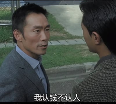 图片中两名男子在户外场景中面对面交谈，左侧男子穿着灰色西装，右侧男子穿着深色纹理外套。背景可见草地、人行道和铁丝网围栏，整体氛围偏向写实。画面底部有白色字幕：“我认钱不认人”，展现出一种直接、现实的对话场景。