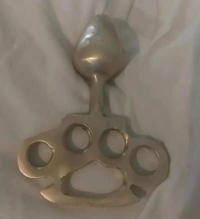 这是一件经过手工改装的金属防身格斗器具，主体保留了传统指虎的结构，拥有四个供手指穿入的圆形孔位，下方还有一个专为拇指设计的特殊形状放置孔，整体材质为金属，表面呈现出金属原色质感，带有制作或使用留下的细微痕迹。在指虎的上方额外加装了一个类似勺子头部的锤头状结构，让这件器具同时具备指虎的握击伤害能力和锤头的砸击伤害能力，属于自制的多功能防身格斗类器具。