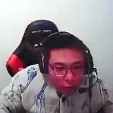 图片中显示一位戴着黑色耳机和眼镜的人，正坐在一把黑色为主、带有红色细节的电竞椅上。人物穿着浅色带有花纹的上衣，背景较为简单，主要聚焦于人物上半身及电竞椅。