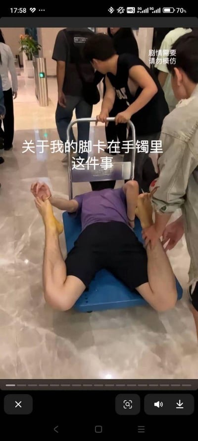 这是一张竖屏短视频截图，画面呈现出一个荒诞搞笑的场景：一名身穿紫色上衣、黑色短裤的男子趴在蓝色平板推车上，他的一只脚和自己的手被一个手镯紧紧卡在一起，无法分开。身旁的几位同伴有的扶着他的脚，有的推着推车，似乎在想办法帮忙。图中配有显眼的文字“关于我的脚卡在手镯里这件事”，右上角还有“剧情需要 请勿模仿”的提示文字，整体是刻意设计的离谱整活情节，通过反常的状况制造出滑稽的社死感，博人一笑。