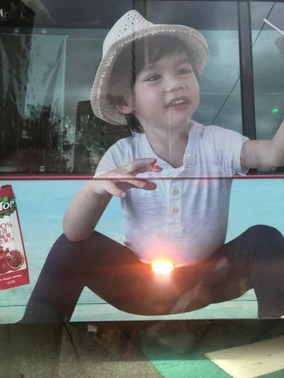 这是一张通过错位拍摄制造趣味效果的图片，广告牌上的外国萌娃穿着白T恤和深色裤子，盘腿坐着并抬手做出姿势，下方的灯光刚好落在他的腹部位置，结合手部动作，看起来像是小孩从身体发出光芒，仿佛在施展超能力，画面左侧还有一盒标注“100%石榴复合果汁”的Top品牌饮料广告。
这是典型的视觉错位梗，利用环境灯光与广告牌的位置巧合，营造出滑稽的视觉错觉，充满搞笑感。