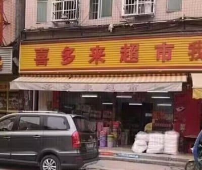 照片展示了一家名为“喜多来超市”的临街店铺外观。黄色招牌上用红色大字书写店名，下方配有白色遮阳篷，门口堆放着白色袋装货物，一辆灰色面包车停在店前街道。背景为多层居民楼，窗户装有防盗网，整体呈现典型的城市社区便利店场景，画面真实自然。