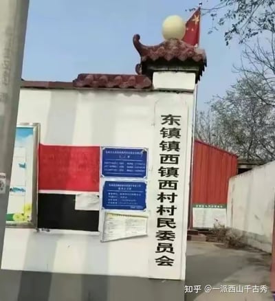 这是一张乡村基层办公地点的实拍照片，画面主体是一块白色立柱式的村委会标识牌，立柱顶部带有中式瓦片造型装饰和圆球，旁侧插有一面中国国旗。立柱上的文字为“东镇镇西镇西村村民委员会”，名称重复了“镇西”字样，读起来绕口易混淆，是这张图的趣味点。标识牌旁的墙面上贴有红黑配色的告示板和几块蓝色通知牌，背景可见树木与红色简易板房，整体呈现出典型的乡村办公场景。