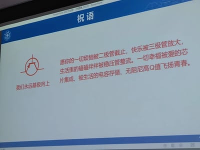 一张PowerPoint幻灯片截图，标题为"祝语"，内容是结合电子工程专业术语创作的祝福语录。幻灯片左上角有一个蓝色校徽或机构标志，中间偏左有一个红色的三极管(或类似电子元件)示意图。主要文字内容使用红色字体，将人生祝福与电子元件功能巧妙结合，如"烦恼被二极管截止"、"快乐被三极管放大"等，体现了理工科特色的创意祝福。底部有"我们永远基极向上"的标语，巧妙运用了电子术语"基极"与"积极向上"的谐音双关。
