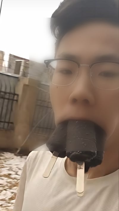 这是一张真人实拍的竖版静态照片，画面主体是一名佩戴半框眼镜、身穿白色上衣的东亚年轻男子，他身处室外场景，地面覆盖有积雪，背景可见建筑物外墙与围栏。该男子将三根巧克力脆皮冰棍同时全部含在嘴里，冰棍的棍身露在嘴外，呈现出搞怪的趣味挑战效果。