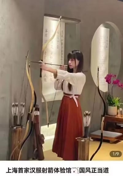 图中一位身着上白下红制式汉服的年轻女孩，正身处充满国风氛围的室内射箭场馆中，双手拉开传统弓箭，眼神专注地瞄准前方。场馆内布置尽显雅致：墙面悬挂着写有汉字的书法卷轴，一旁的木质箭筒中插满箭矢，角落还摆放着一盆盛开的兰花，整体环境与汉服、射箭的传统元素相得益彰。图片下方配有文字“上海首家汉服射箭体验馆 🎋国风正当道”，突出了这是将传统汉服文化与射箭运动结合的新式体验场馆，展现出国风文化当下流行的趋势。