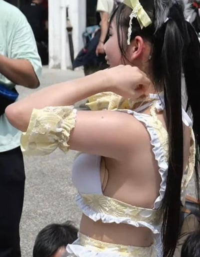 这是一张户外拍摄的照片，画面主体是一位扎着黑色双马尾、头戴黄色蝴蝶结发饰的女生，她穿着黄白配色的可爱风格cos服装，侧过身，一只手抬起放在脖颈附近，似乎在整理衣物或是摆拍。背景中能看到模糊的其他路人，场景看起来像是在漫展或者类似的线下ACG活动现场，整体氛围轻松活泼，充满了二次元活动的氛围感。
