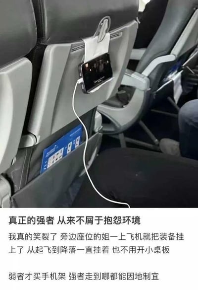 图片展示了飞机座椅后背的创意用法：一位乘客将手机巧妙地固定在座椅后背的纸巾盒上作为临时支架，手机连接着充电线，无需使用小桌板即可舒适观影。图片下方配有文字："真正的强者 从来不屑于抱怨环境 我真的笑裂了 旁边座位的姐一上飞机就把装备挂上了 从起飞到降落一直挂着 也不用开小桌板 弱者才买手机架 强者走到哪都能因地制宜"，赞扬这种因地制宜的生活智慧。