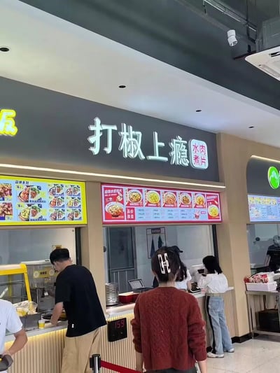 这是一张在美食城或食堂拍摄的快餐门店实拍图，门店招牌为「打椒上瘾」，主打水煮肉片品类。招牌的「打椒上瘾」是谐音梗，谐音「打胶上瘾」，以此突出菜品麻辣口感让人上头的特点。柜台上方设有电子菜单，左侧展示双拼系列餐品（价格多为16元），中间展示水煮肉片系列餐品（含麻辣、金汤酸菜等口味），柜台前有多位顾客正在点餐或取餐，门店装修为现代简约的快餐档口风格。