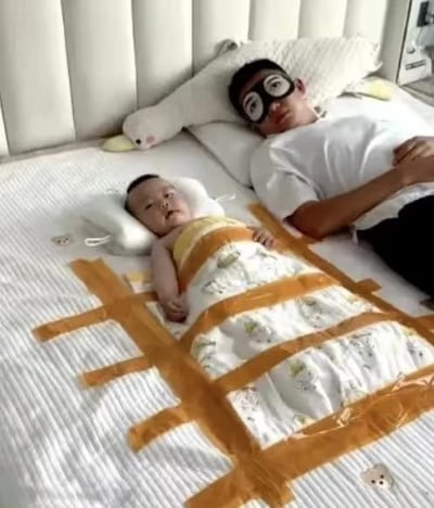 图片中，一名男子戴着黑色眼罩躺在床上，身旁是一个被黄色胶带以网格状固定在床铺上的婴儿。婴儿裹着印有卡通图案的白色毯子，头部枕着白色小枕头，表情平静。男子穿着白色T恤和黑色裤子，姿态放松。床铺上铺着白色条纹床单，背景有白色床头板和一个白色毛绒玩具。整个画面展现了一种幽默又温馨的亲子日常场景，父亲用胶带固定宝宝防止其乱动，是一种有趣的育儿方式。