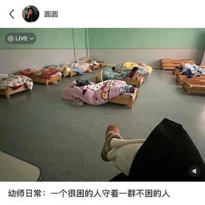 这是一张直播画面截图，呈现了幼儿园午睡室的场景：室内摆放着多张小木床，床上盖着花色各异被子的小朋友们，有的并未入睡还保持着活跃状态；画面下方露出拍摄者（一名幼师）的腿，看起来正处于放松的状态。画面底部配文“幼师日常：一个很困的人守着一群不困的人”，精准吐槽了幼师在午睡时段的无奈日常——自己困意十足，却要照看精力旺盛不肯午睡的小朋友，内容真实接地气，能引发幼师群体的强烈共鸣，充满了搞笑又心酸的氛围。