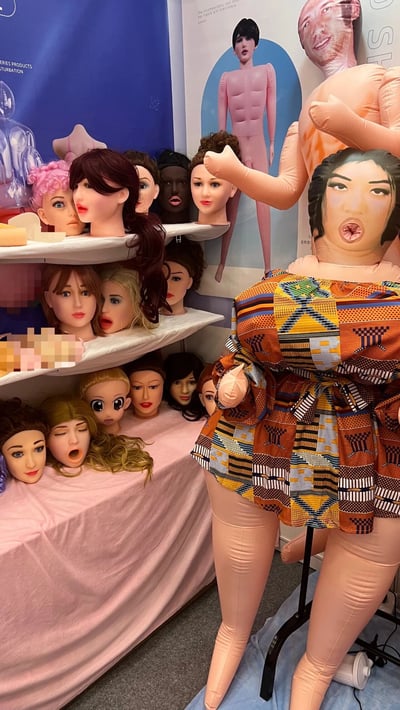 图片展示了一个奇特的商品展示场景，左侧多层架子上整齐摆放着多个不同发型、发色和表情的人形模特头，部分模特头面部被像素化处理（可能为内容合规）。右侧有一个大型充气人形玩偶，身着色彩鲜艳的几何图案服装，姿态夸张。背景中可见印有充气人形模特的海报，整体场景呈现出一种怪异且略带荒诞的视觉效果。