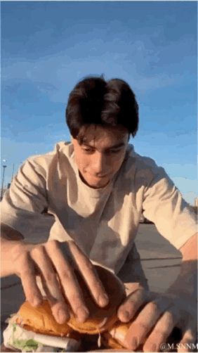 这是一段趣味整蛊类动态GIF，画面中一名男生在户外场景下，双手按住汉堡的上下面包，通过视觉错位让观众以为这是一个巨型汉堡；当他拿开双手后，真相揭晓——其实是多个小汉堡叠在一起只露出上下层面包，制造出有趣的反转效果，让人忍俊不禁。