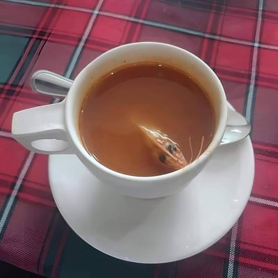 一张实拍的趣味图片，白色带把手的马克杯中装着类似港式奶茶的棕色饮品，饮品里居然浸泡着一个完整的带虾须的虾头，杯子放置在配套的白色杯碟上，杯碟旁搭着一把金属勺子，背景是红黑格子图案的桌布。这种完全不搭的食材组合，像是餐厅出餐失误或是网友整活的黑暗料理，画面充满离谱感，让人忍俊不禁。