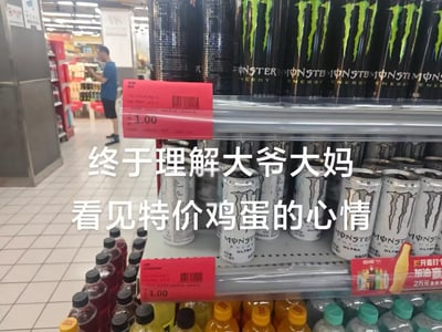这是一张在超市内拍摄的实拍梗图，画面主体是超市的饮料货架，货架上整齐陈列着黑罐、白罐两款魔爪能量饮料，对应位置的红色价签清晰标注售价为1元，远低于该商品的常规市场价。图中叠加了醒目的白色大字配文，将年轻人看到喜欢的饮料超低价促销时的惊喜、想要抢购的心情，和老年群体看到特价鸡蛋时的心态做类比，充满趣味性和共鸣感，背景处还能看到超市的公共过道，以及一名正在远处货架旁挑选商品的男性顾客。