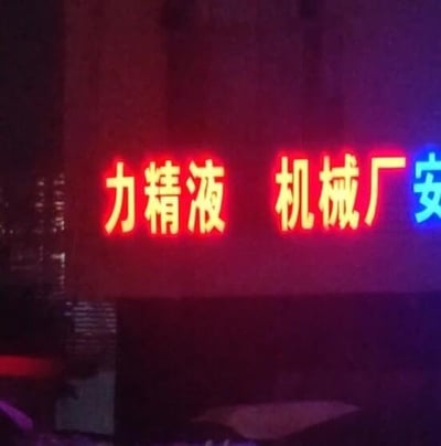 图片展示了夜晚场景下一栋建筑物外墙上的霓虹灯招牌。招牌主体为红色发光字体，左侧显示“力精液”，中间为“机械厂”，右侧可见部分蓝色字体“安”（推测为“安全”等词语的不完整显示）。由于“力精液”一词的组合易引发不雅联想，属于因文字排列或可能的错别字导致的搞笑场景。