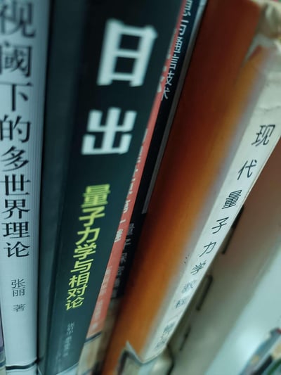 这是一张近距离拍摄的书架上的书籍书脊照片，展示了多本物理相关的专业书籍，包括《视阈下的多世界理论》《日出 量子力学与相对论》《现代量子力学教程（修订版）》，还有一本部分被遮挡的信息通信技术类书籍，书籍排列紧密，呈现出专业的物理学科阅读收藏的场景。图中无谐音梗、双关或暗示类内容。