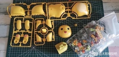 绿色切割垫上整齐摆放着黄色塑料模型套件的零件，包含标有'A2'的多组流道（sprues），上面连接着各种模型部件。中央位置有一个已从流道上取下的黄色角色头部，面部带有微笑表情，显得十分治愈。旁边散落着小型黄色部件和一个密封透明塑料袋，内含多种彩色小零件。右下角可见水印'@大班模型吧'，整体场景展现了模型制作前的零件准备状态，适合模型爱好者分享交流。