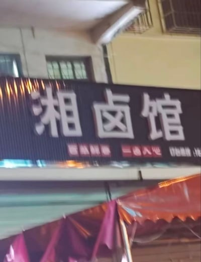 这是一张竖版实拍照片，展示了一家名为“湘卤馆”的餐馆招牌。招牌主体为深色背景，上方有醒目的白色大字“湘卤馆”，下方隐约可见红色和白色的小字，推测为宣传语或联系方式。背景中能看到老旧居民楼的部分结构，包括带铁栅栏的窗户和外挂空调外机，招牌下方有红色的遮阳棚。照片整体略显模糊，呈现出街头随拍的真实感。
