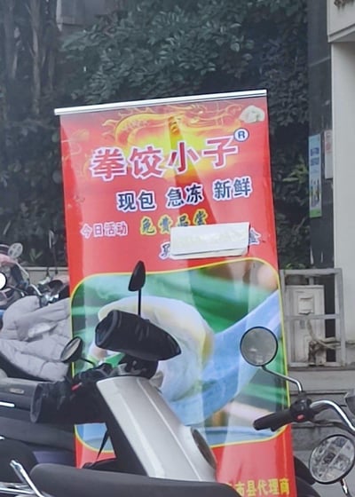这是一张户外实拍的餐饮宣传展架照片，展架以鲜艳的红色为底色，是速冻饺子品牌“拳饺小子”的线下推广物料，主打“现包 急冻 新鲜”的产品卖点，当日推出“免费品尝”的引流活动，展架下方配有饺子的实物展示图。展架被前景的电动车部分遮挡，背景可见绿植、停放的其他电动车以及街边建筑设施，整体呈现出街边餐饮商家线下引流的日常场景。