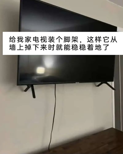 这是一张静态实拍图，画面主体是一台挂在墙面上的壁挂式电视，电视的下方安装了原本设计用于放置在桌面的电视脚架。图片上方配有白色背景的黑色文字：“给我家电视装个脚架，这样它从墙上掉下来时就能稳稳着地了”。这是一种无厘头的迷惑操作，脚架在电视正常挂墙时无法发挥支撑作用，只有在电视意外掉落的场景下才可能让电视“稳稳着地”，通过这种荒诞的脑洞制造出搞笑的效果，展现了一种趣味的生活恶搞行为。
