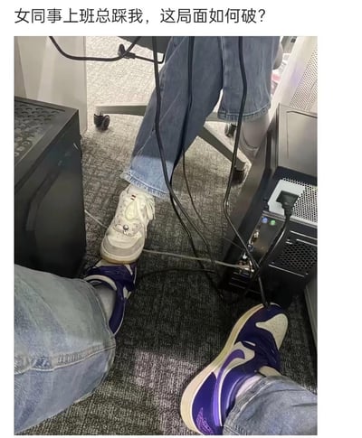 图片内容为办公室场景中两个人的脚交叠在一起，一人穿紫色 Nike 球鞋，一人穿休闲鞋，画面中出现了办公桌下的电脑主机、电线等。配文为“女同事上班总踩我，这局面如何破？”，表现职场中人与人之间的空间尴尬及搞笑局面。整个场景具有一定的生活感和幽默色彩，经常被用来表达职场中的无奈或好玩的时刻。
