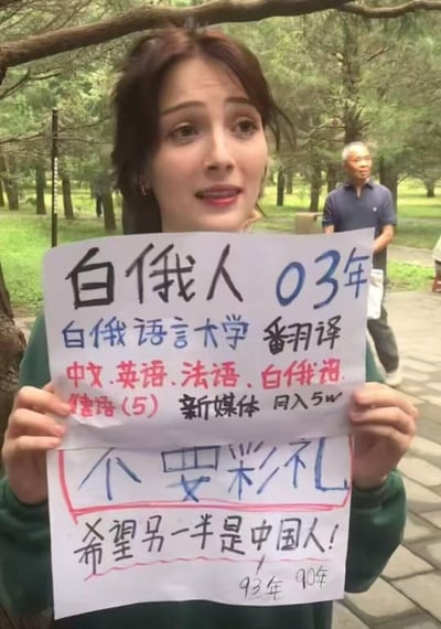 图片中一位年轻女子在公园环境中手持手写纸牌，纸牌上用中文详细写着个人信息：白俄人，2003年出生，白俄语言大学翻译专业，掌握中文、英语、法语、白俄语和德语（5级），从事新媒体工作月入5万元。最引人注目的是纸牌下方特别标注“不要彩礼”，并明确表示“希望另一半是中国人”，还写有“93年 90年”可能是对另一半年龄的期望。背景中可以看到公园的树木和一位路过的老人，整体氛围自然真实。