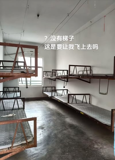 一间宿舍房间内摆放着多组双层床，其中部分上铺直接固定在较高的墙壁上，但没有配备任何梯子或爬梯装置。图片上方配有文字"? 没有梯子 这是要让我飞上去吗"，以幽默的方式吐槽这种不合理的床铺设计。房间整体较为简陋，水泥地面，墙壁上可见裸露的电线，靠窗位置有自然光射入。