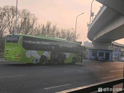 这是一张在城市路口拍摄的实拍照片，画面主体是一辆京A牌照的绿色北京新能源公交车，车身在中部位置发生明显断裂，前后车身分离，一名人员站在断裂的车身旁，似乎在查看事故情况。背景可见城市高架桥、路边的简易蓝色小屋、光秃的树木，天色呈现黄昏时分的暖橙色，路面为城市柏油马路，整体场景显示这是一起公交车在运营过程中发生的意外故障事故。