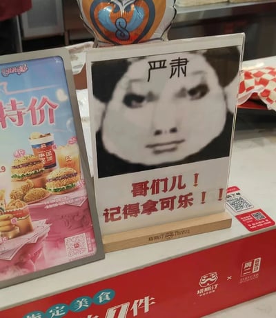 这张图片拍摄于塔斯汀中国汉堡的线下门店取餐区域，画面主体是两个立牌和取餐台的部分区域。右侧的立牌是一个趣味提示牌，上方是被P上“严肃”字样的熊猫头搞笑表情包，表情包呈现出一本正经的神态，下方用加粗的红色字体写着“哥们儿！记得拿可乐！！”，用接地气的网络风格话术提醒取餐的顾客不要遗漏配套的可乐；左侧是塔斯汀的特价套餐宣传立牌，展示了包含迷你中国堡、小食和饮品的组合套餐，标注了价格和套餐内容；取餐台的右侧还有扫码下单的二维码，台面上有塔斯汀的品牌标识，整体场景充满了轻松幽默的氛围，用年轻人喜欢的搞笑方式传递提示信息，拉近了品牌和顾客的距离。