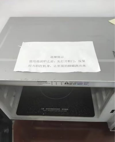 这是一张实拍的公共使用微波炉的照片，银灰色微波炉的顶部放置了一张打印的白色提示纸，纸上写着温馨提示：使用微波炉之前，先打开柜门，反复用力拍打机身，让里面的蟑螂跑出来。该提示直白说明该微波炉内有蟑螂出没的问题，内容既实用又带有荒诞的搞笑感，反映了部分公共区域家电的卫生状况。