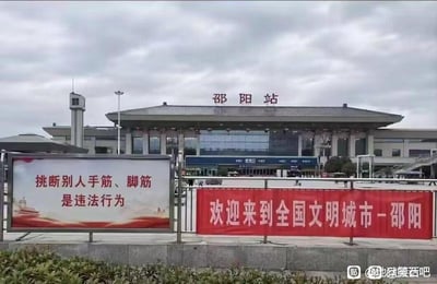 这是一张拍摄于邵阳站站前的实拍图，背景是阴天状态下的邵阳站站房建筑，站房顶部标有“邵阳站”字样。前景护栏上并列设置了两块宣传物料：左侧是白底宣传板，用红色文字写着“挑断别人手筋、脚筋是违法行为”；右侧是红色横幅，用白色文字写着“欢迎来到全国文明城市－邵阳”，二者内容并置形成了强烈的反差感，图片右下角带有百度贴吧“地狱笑话吧”的水印。