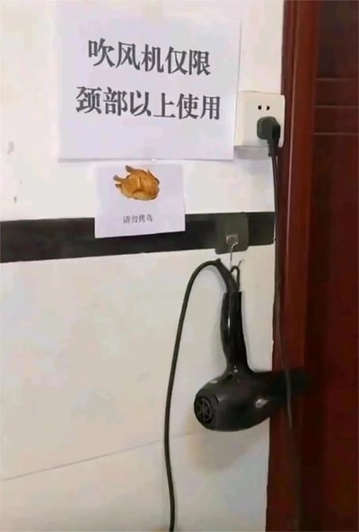 这是一张竖屏实拍的浴室场景图，墙面为白色瓷砖，右侧可见深色木质门框。墙面上张贴着两张趣味提示标语：上方白色纸张写有“吹风机仅限颈部以上使用”，下方小纸张印有烤鸡图案，配文“请勿烤鸡”，两者之间用黑色胶带做了视觉分隔。一个黑色吹风机挂在墙面上的挂钩上，其插头插在右侧的白色墙壁插座中。这组标语以幽默的方式，提醒使用者吹风机只能用于吹干头发（颈部以上部位），禁止用其进行加热食物等违规、危险的操作，常见于公共浴室、集体宿舍这类需要规范电器使用的场所。