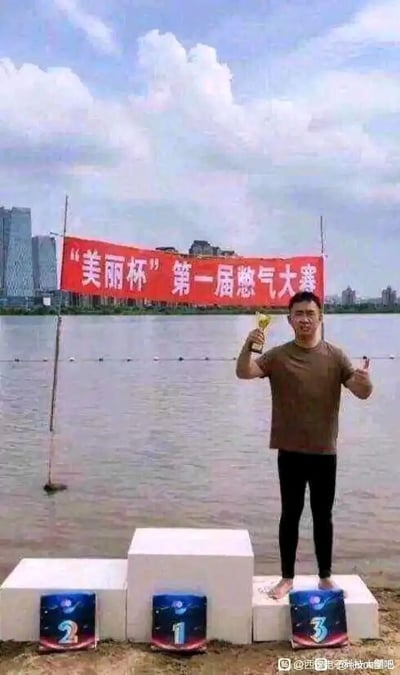 这是一张在湖边拍摄的恶搞趣味图，背景是开阔的水面与远处的城市建筑群，岸边的水面上方悬挂着一条红色横幅，上面写着“‘美丽杯’第一届憋气大赛”。岸边设置了三个白色的颁奖台，从左到右的台子下方分别贴着名次标识2、1、3，其中1号台子高度最高，2号次之，3号最矮，与常规颁奖台的名次高度对应逻辑一致，但标识的位置却和常规认知相反。一名赤脚的男子站在最右侧的3号矮台子上，他身着棕色短袖上衣与黑色长裤，右手举着奖杯，双手做出庆祝的手势，脸上带着兴奋的表情。这张图的幽默点在于结合“憋气大赛”的主题，暗示其他参赛选手可能还在水下憋气未上岸，只有这名男子完成比赛站在台上，同时颁奖台的名次标识与人物位置形成错位反差，制造出荒诞搞笑的效果。