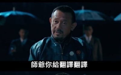 电影《让子弹飞》中的夜晚场景，前景中主角张麻子（姜文饰）身着深色中式外套，表情严肃地站立，背景有几位撑着雨伞的人物伫立。画面底部配有标志性白色字幕“师爺你給翻譯翻譯”，该台词因电影情节中对“翻译”行为的反复强调而成为经典网络梗，整体氛围庄重且带有戏剧张力。