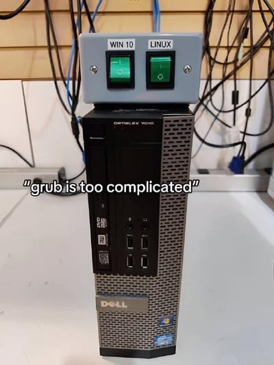 图片展示了一台戴尔Optiplex 7010电脑主机，其顶部安装了一个自制的物理切换装置。该装置有两个绿色按钮，分别标记为"WIN 10"和"LINUX"，用于直接切换电脑启动的操作系统。背景中可以看到许多杂乱的连接线。图片下方配有文字"grub is too complicated"，暗示这种物理切换方式是为了避免使用复杂的GRUB引导程序。这是一种幽默且实用的技术解决方案，体现了程序员面对复杂软件时的创意应对方法。