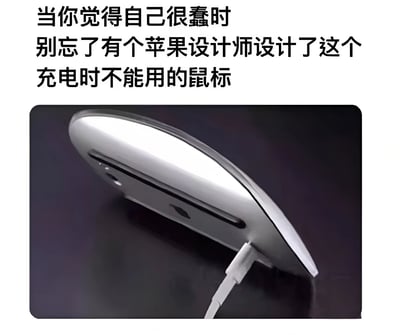 这张梗图嘲讽了苹果Magic Mouse的设计缺陷——充电接口位于鼠标底部，导致充电时无法使用。图片展示了银色的鼠标底部插着充电线，上方文字写道：‘当你觉得自己很蠢时，别忘了有个苹果设计师设计了这个充电时不能用的鼠标。’
