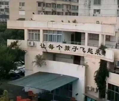 一张拍摄于城市住宅区的照片，画面中央是一栋略显陈旧的白色建筑，建筑外墙上有黑色大字标语“让每个孩子飞起来”。建筑周围可以看到其他居民楼，楼下停有车辆，部分墙面有植物攀爬生长，展现出生活化的场景氛围。