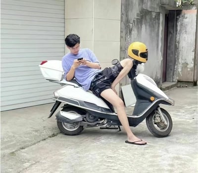 图片中一名少年身穿休闲装扮，慵懒地躺在摩托车上刷手机，姿势十分随意松弛。他的腿跨在摩托车边上，旁边把手上摆放着黄色头盔，远看像人头直接长在车前，形成了一个令人误会的视觉错位效果。整体氛围带着幽默和轻松，比较接近网络上的佛系或“人在魂在”这类错位趣味梗。

特殊提示：头盔与身体错位，制造“人在魂在”、“魂穿机车”等视觉笑点。
