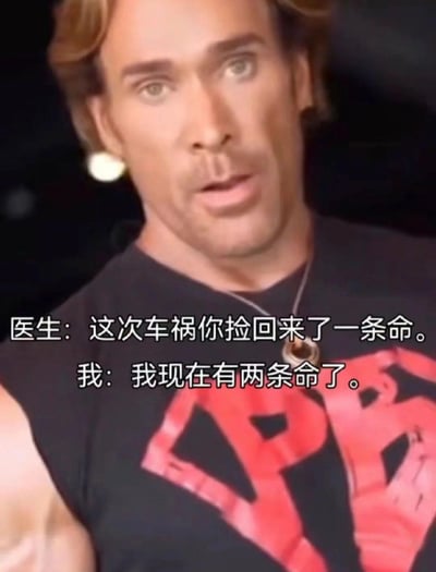 图片中是美国健身网红迈克·奥赫恩，他穿着黑色无袖背心，上面有红色图案，表情展现出典型的"迷之自信"。图中配有对话文字："医生：这次车祸你捡回来了一条命。我：我现在有两条命了。" 这是典型的"大力王文学"表现形式，通过无逻辑的对话来讽刺盲目自信的态度。