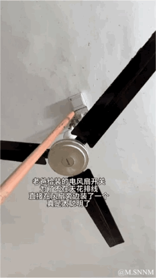 这张图展示了一个安装在吊扇电机侧面的开关，还有一根木棍伸向开关似乎要去按压它。图中配文吐槽老爸为了不在天花板排线，直接把风扇开关装在了风扇旁边，这个操作看似“聪明”实则非常不合理——风扇运转时根本无法触碰开关，即便风扇静止，用木棍去按也十分不便，充满了生活里的搞笑反差感，让人哭笑不得。